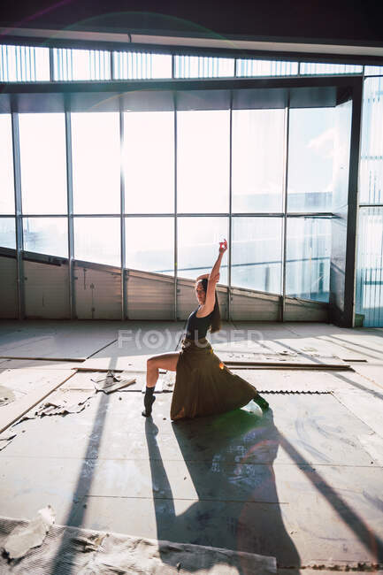 Side view of female theater artist dancing with raised arm looking at camera on floor with shadow against glass wall — Stock Photo
