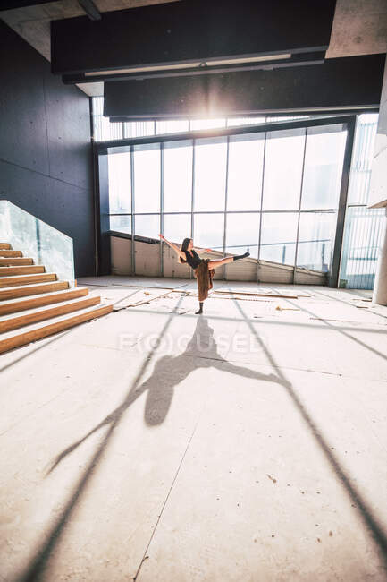 Side view of unrecognizable female theater artist dancing with raised leg and arm on floor with shadow against glass wall — Stock Photo