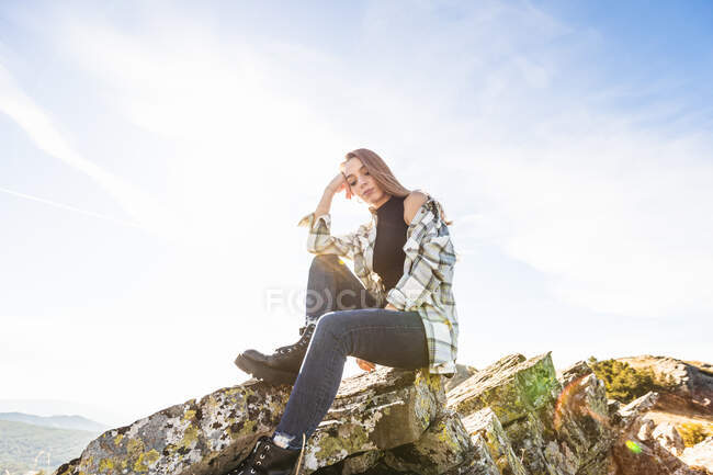 From below of young melancholic female traveler in casual wear and boots leaning on hand while sitting on mount and looking down — Stock Photo