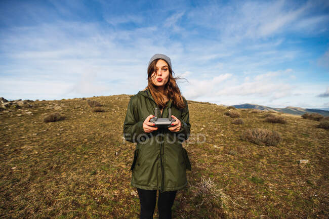 Молодая туристка с надутыми губами, управляющая беспилотником с дистанционным управлением, глядя в камеру на лужайке в Европе — стоковое фото