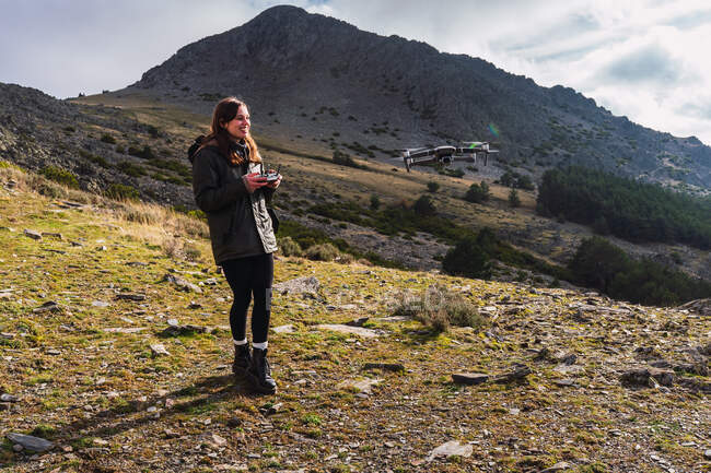 Seitenansicht einer jungen, fröhlichen Touristin, die Drohne mit Fernbedienung steuert, während sie sich auf den Bergrücken freut — Stockfoto