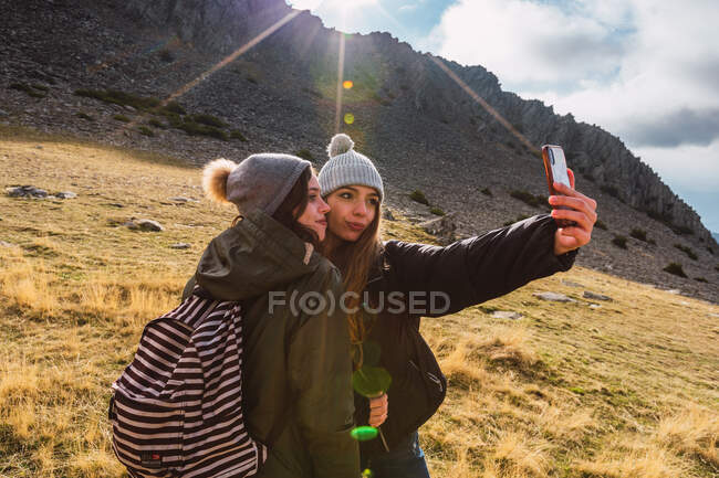 Junge zufriedene Freundinnen mit Rucksäcken machen Selfie mit dem Smartphone, das im Gras vor einem Berg in der Sonne steht — Stockfoto