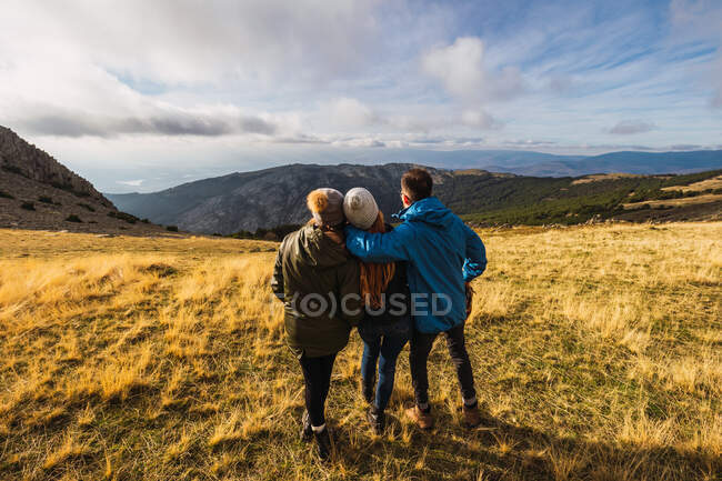 Rückansicht eines anonymen Mannes, der Freundinnen umarmt, während er unter wolkenverhangenem Himmel in Spanien Berge aus dem Rasen betrachtet — Stockfoto