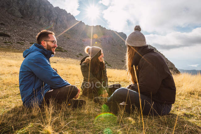 Viaggiatori allegri in capispalla che parlano mentre riposano sull'erba contro cresta sotto il cielo nuvoloso alla luce del sole — Foto stock