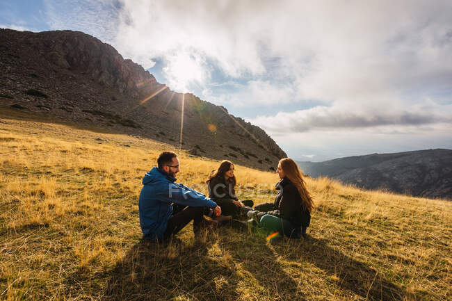Веселих подорожніх, які розмовляють надворі, відпочиваючи на траві під хмарним небом під сонцем. — стокове фото