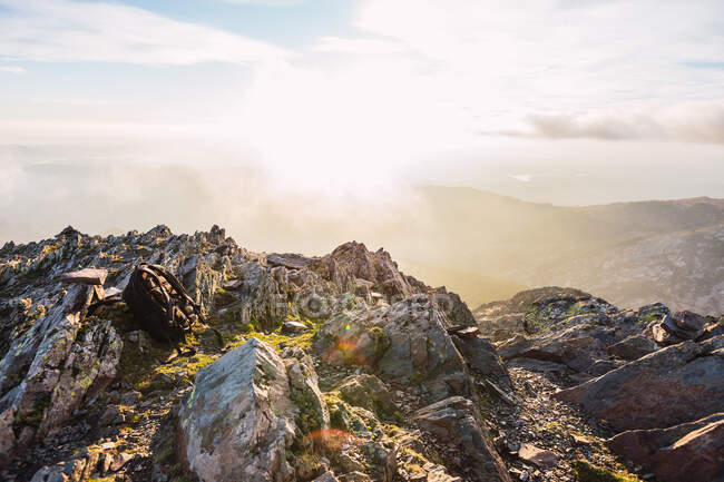 Pacote traseiro descansando em rochas perto de cumes altos com neblina sob o céu azul brilhante com nuvens à luz do sol — Fotografia de Stock