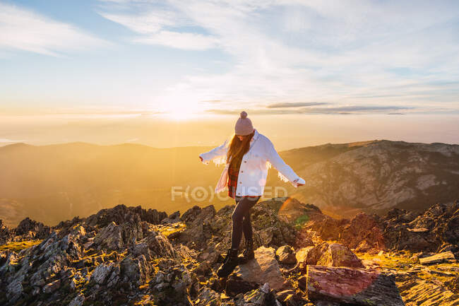 Анонімна жінка-туристка в верхньому одязі, що гуляє по грубих каменях в горах під хмарним небом на блискучому світанку в спині освітленому — стокове фото