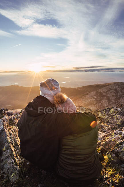 Vue arrière d'un homme méconnaissable embrassant sa petite amie en vêtements de dessus tout en admirant l'aube de la montagne sous un ciel nuageux au soleil — Photo de stock