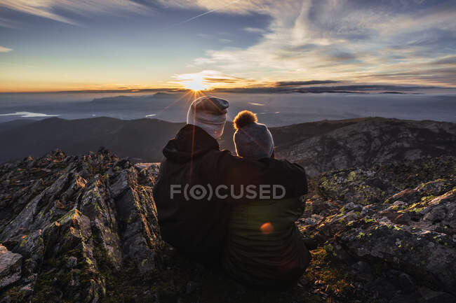 Back view of unrecognizable man embracing girlfriend in outerwear while admiring dawn from mountain under cloudy sky in sunshine — Stock Photo