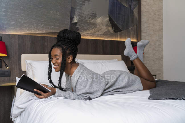 Vista lateral da jovem estudante afro-americana feliz em roupa casual deitada na cama confortável e livro de leitura em casa — Fotografia de Stock