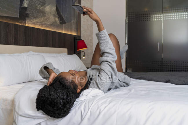 Весела чорна жінка-підліток у теплому светрі та шкарпетках лежить на зручному ліжку та сміється, маючи відеодзвінок на смартфон вдома — стокове фото