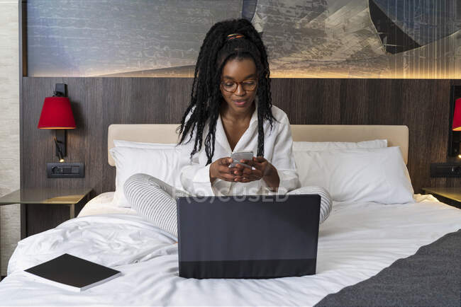 Jeune pigiste afro-américaine concentrée aux cheveux longs bouclés en vêtements décontractés et lunettes assises sur un lit confortable avec ordinateur portable et ordinateur portable et utilisant un smartphone pendant le travail à distance — Photo de stock