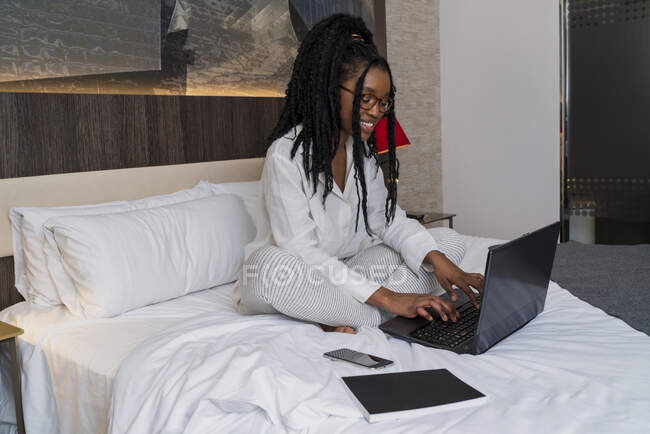 Concentrated young African American female freelancer with curly long hair in casual clothes and eyeglasses sitting on comfortable bed with laptop and notebook during remote work — Stock Photo