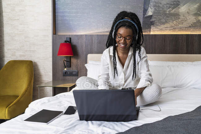 Joven estudiante afroamericana positiva con ropa casual y auriculares sonriendo mientras está sentada en el acogedor be y escribiendo en la computadora portátil durante la lección en línea en casa - foto de stock