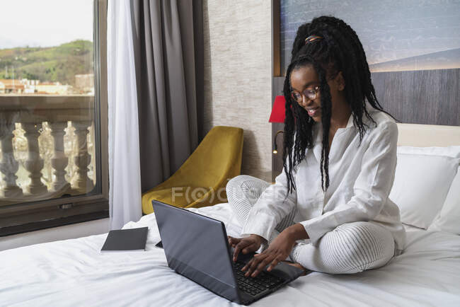 Concentrada joven afroamericana freelancer con pelo largo rizado en ropa casual y anteojos sentados en cómoda cama con portátil y portátil durante el trabajo remoto - foto de stock