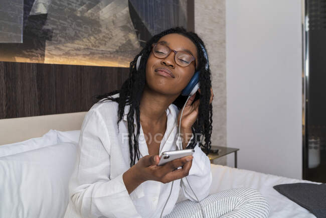 Joven mujer afroamericana positiva en ropa casual y auriculares sonriendo con los ojos cerrados sentados en la acogedora cama con teléfono inteligente en casa - foto de stock