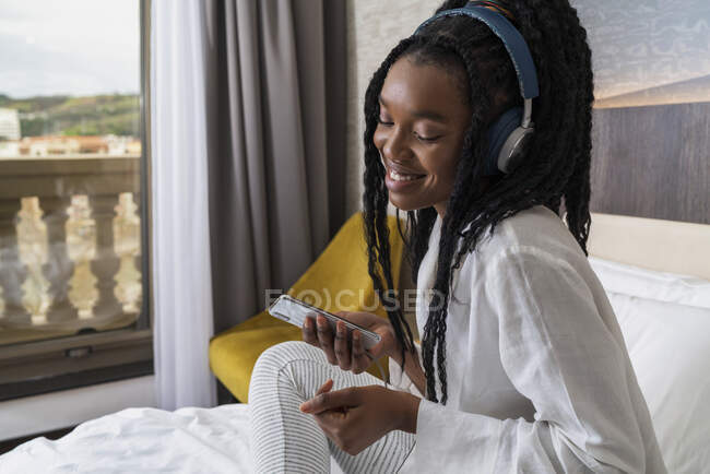 Side view of positive young African American female in casual clothes and headphones smiling while sitting on cozy bed with smartphone at home — Stock Photo