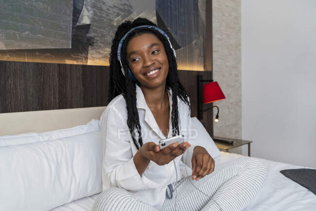 Positive jeune afro-américaine femme en vêtements décontractés et écouteurs souriant tout en étant assis sur un lit confortable avec smartphone à la maison — Photo de stock