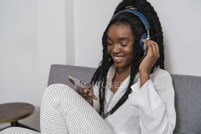 Joven dama afroamericana alegre con el pelo largo rizado en ropa casual sonriendo mientras escucha música en los auriculares y mensajes en el teléfono inteligente sentado en el sofá en casa - foto de stock