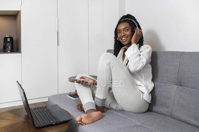 Радісна молода афроамериканська леді з довгим кучерявим волоссям в повсякденному одязі посміхається, слухаючи музику в навушниках з смартфоном і працюючи на комп'ютері, сидячи на дивані вдома. — стокове фото