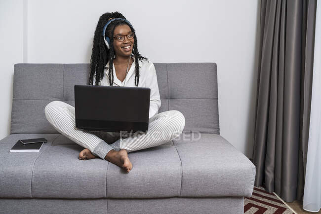 Орієнтована молода етнічна фрілансерка з довгим афро волоссям в повсякденному одязі та окулярах, що працюють дистанційно на ноутбуці та слухають музику в навушниках в сучасній квартирі — стокове фото