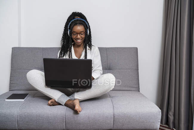 Giovane freelance etnico femminile focalizzato con lunghi capelli afro in abiti casual e occhiali che lavorano da remoto sul laptop e ascoltano musica in cuffia in appartamento moderno — Foto stock