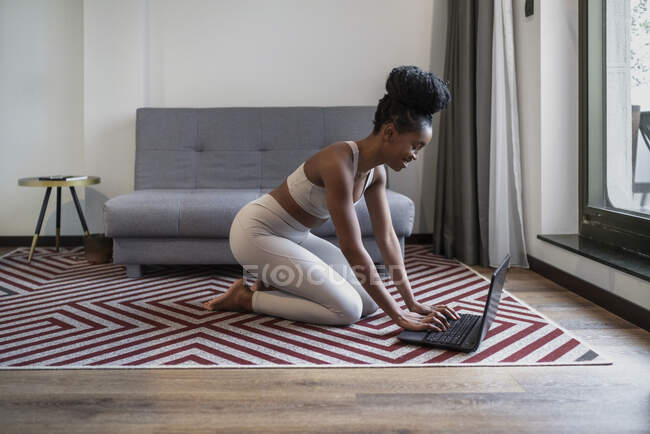 Vista lateral de la joven mujer afroamericana feliz en ropa deportiva sonriendo mientras usa el portátil sentado en el suelo antes del entrenamiento de yoga en línea - foto de stock