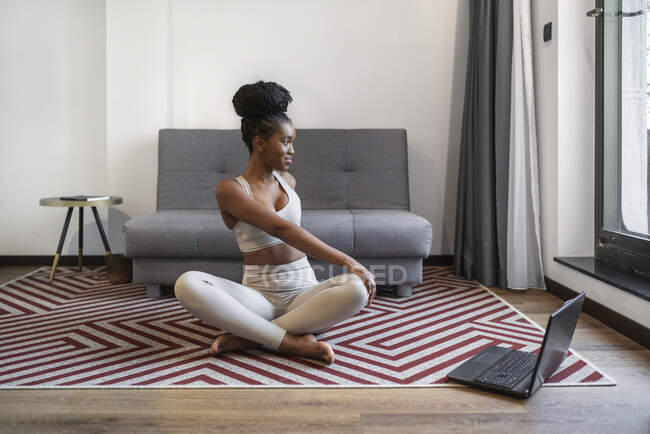 Corps entier de la jeune femme noire concentrée en vêtements de sport assis sur le tapis regarder la vidéo sur ordinateur portable et effectuer la pose de yoga pendant l'entraînement de yoga à distance à la maison — Photo de stock