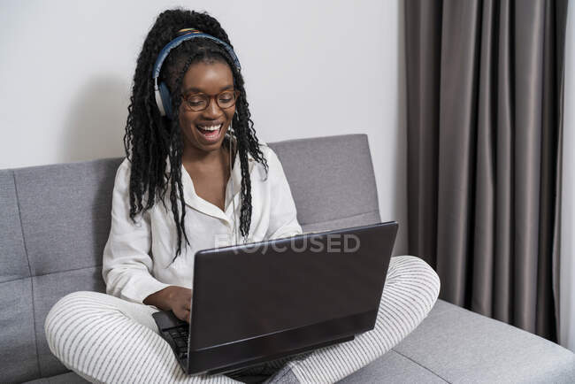 Joven freelancer étnica enfocada con pelo largo Afro en ropa casual y anteojos trabajando remotamente en laptop y escuchando música en auriculares en apartamento moderno - foto de stock