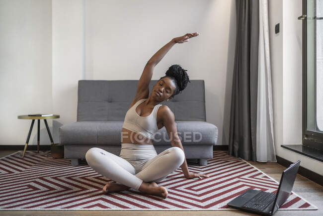 Полное тело концентрированной молодой черной женщины в активной форме сидя на циновке смотреть видео на ноутбуке и выполняя йогу позу во время дистанционного обучения йоге дома — стоковое фото