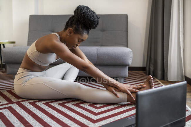 Vue latérale complète du corps de la jeune femme noire concentrée en vêtements de sport assis sur le tapis regarder la vidéo sur ordinateur portable et effectuer la pose de yoga pendant l'entraînement de yoga à distance à la maison — Photo de stock