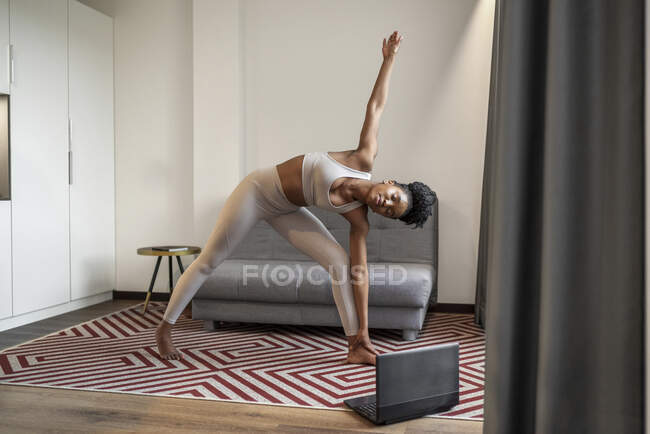Corps complet de jeune femme noire concentrée en vêtements de sport regarder la vidéo sur ordinateur portable et effectuer la pose d'angle latéral pendant l'entraînement de yoga à distance à la maison — Photo de stock