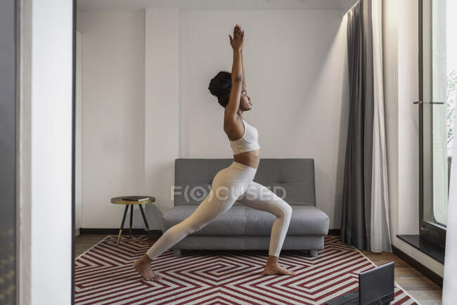 Full body of concentrated young black female in activewear watching video on laptop and performing Virabhadrasana pose during distance yoga training at home — Stock Photo