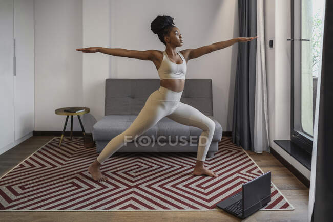Voller Körper einer konzentrierten jungen schwarzen Frau in aktiver Kleidung, die Videos auf dem Laptop anschaut und Virabhadrasana-Posen während des Fernyoga-Trainings zu Hause ausführt — Stockfoto