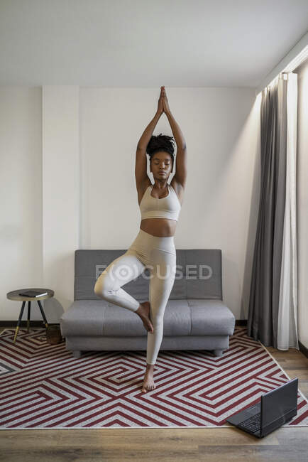 Cuerpo completo de joven concentrada negra en ropa deportiva viendo vídeo en portátil y realizando pose Vrikshasana durante el entrenamiento de yoga a distancia en casa - foto de stock