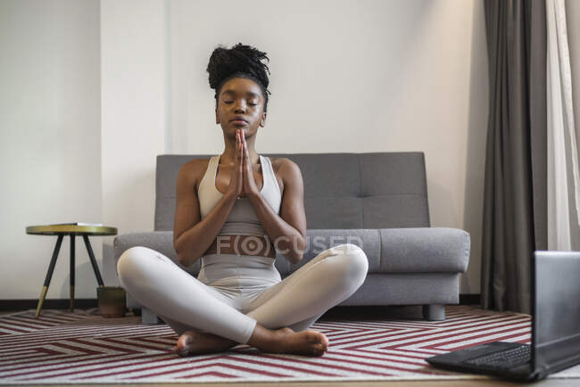 Corpo pieno di rilassato giovane donna afroamericana in abbigliamento sportivo meditando in posa Lotus con gli occhi chiusi e le mani di preghiera durante la sessione di yoga online a casa — Foto stock