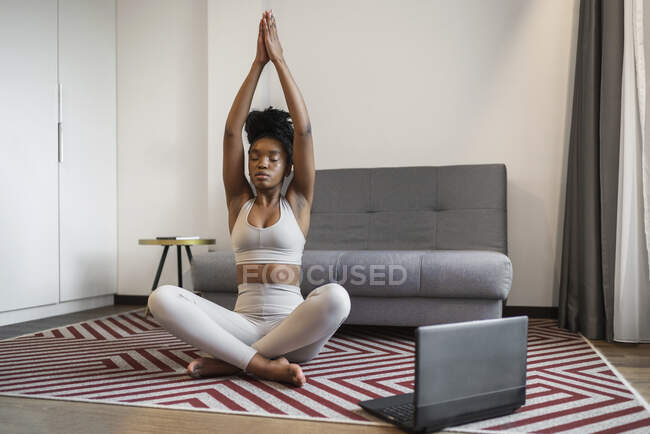 Full body of relaxed young African American woman in sportswear meditating in Lotus pose with closed eyes and prayer hands over head during online yoga session at home — Stock Photo