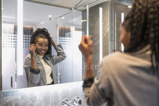 Back view of crop self assured happy young black female millennial in trendy outfit smiling and touching long Afro hair while looking in mirror in bathroom — Stock Photo