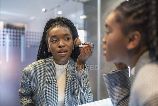 Vista lateral da cultura confiante jovem senhora afro-americana com cabelos longos e escuros olhando no espelho e aplicando pó no rosto com escova no banheiro — Fotografia de Stock
