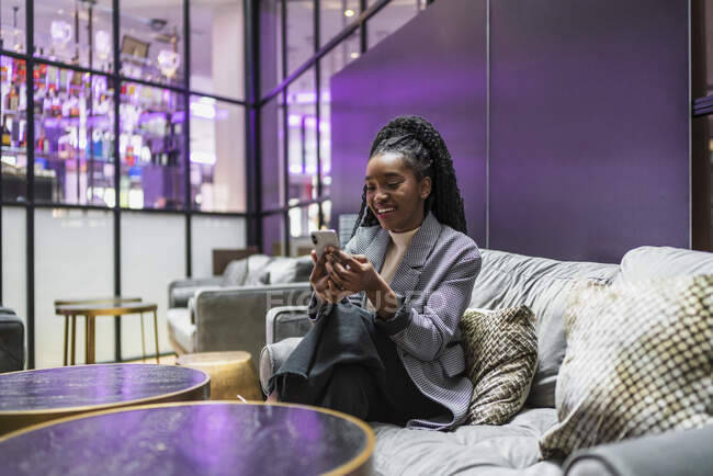 Feliz joven afroamericana femenina con el pelo largo y rizado en traje de moda sentado en cómodo sofá en la cafetería moderna, mientras que los mensajes de texto en el teléfono inteligente - foto de stock