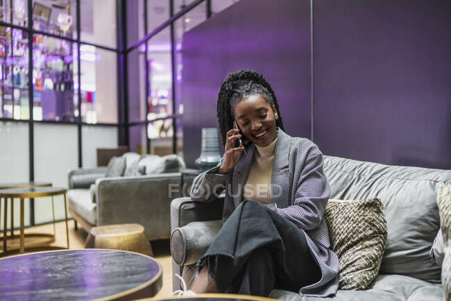 Feliz joven afroamericana femenina con el pelo largo rizado en traje de moda sentado en cómodo sofá en la cafetería moderna y tener conversación telefónica - foto de stock