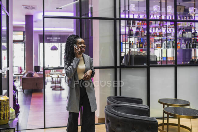 Cheerful trendy young ethnic female millennial with long Afro hair in stylish outfit standing in modern cafe and talking on mobile phone — Stock Photo