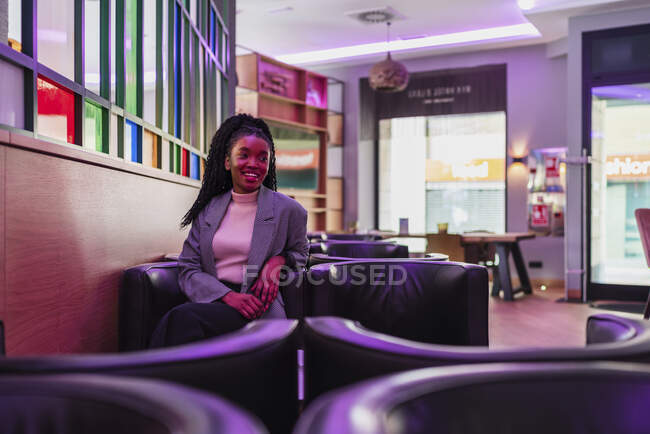 Улыбающаяся молодая афроамериканка тысячелетия с длинными вьющимися волосами в модной одежде, улыбающаяся во время отдыха в удобном кожаном кресле в современном кафе — стоковое фото
