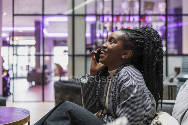 Вид сбоку на счастливую молодую афроамериканку с длинными вьющимися волосами в модном наряде, сидящую на удобном диване в современном кафе и разговаривающую по телефону — стоковое фото