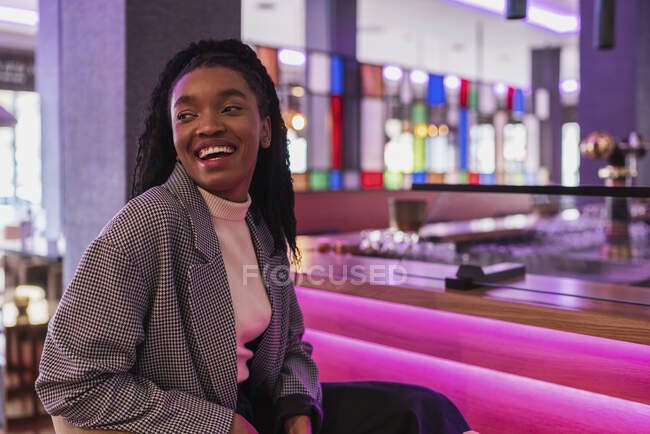 Self assured cheerful young African American lady with long curly hair in stylish outfit smiling while sitting at illuminated counter in modern bar looking away — Stock Photo