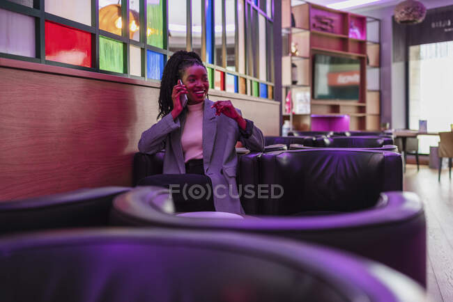 Sorridente giovane afroamericana millenaria con lunghi capelli ricci in abiti alla moda sorridenti mentre riposa in comoda poltrona in pelle in un caffè contemporaneo parlando su smartphone — Foto stock