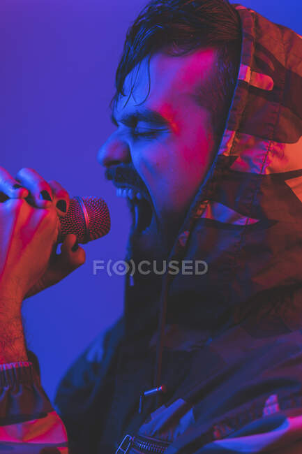 Adult bearded male singer in hooded jacket performing expressive song with microphone during rock concert in neon illumination — Stock Photo