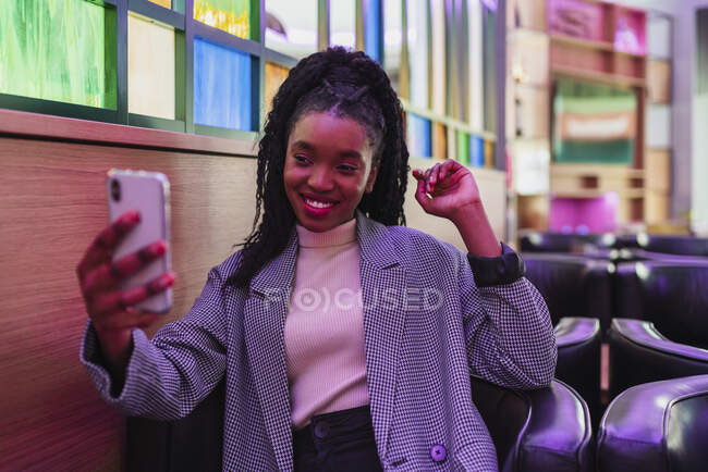 Elegante joven mujer afroamericana con pelo largo y rizado oscuro en traje de moda sonriendo mientras toma selfie en el teléfono inteligente en la cafetería moderna - foto de stock