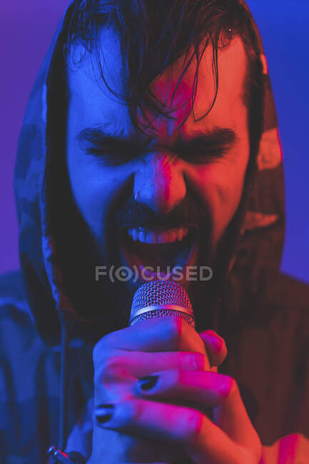 Adult bearded male singer in hooded jacket performing expressive song with microphone during rock concert in neon illumination — Stock Photo