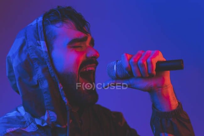Cantante maschio adulto barbuto in giacca incappucciata che esegue canzone espressiva con microfono durante concerto rock in illuminazione al neon — Foto stock